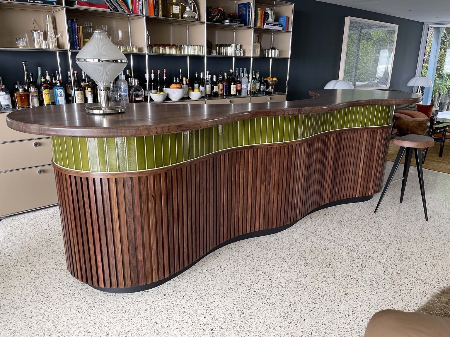 Curved custom home bar with vertical wood slat cladding, a glossy olive-green tile band, and a live-edge wood top on stone flooring