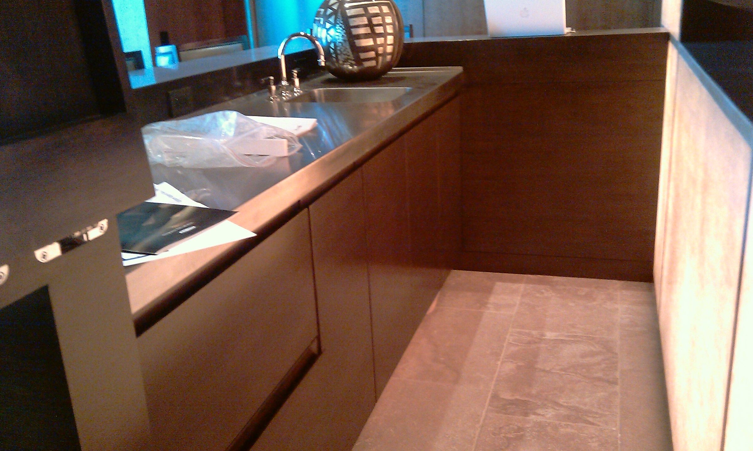 L-shaped custom wood wet bar with a stainless double-basin sink, gooseneck faucet, mirrored backsplash, and stone tile flooring