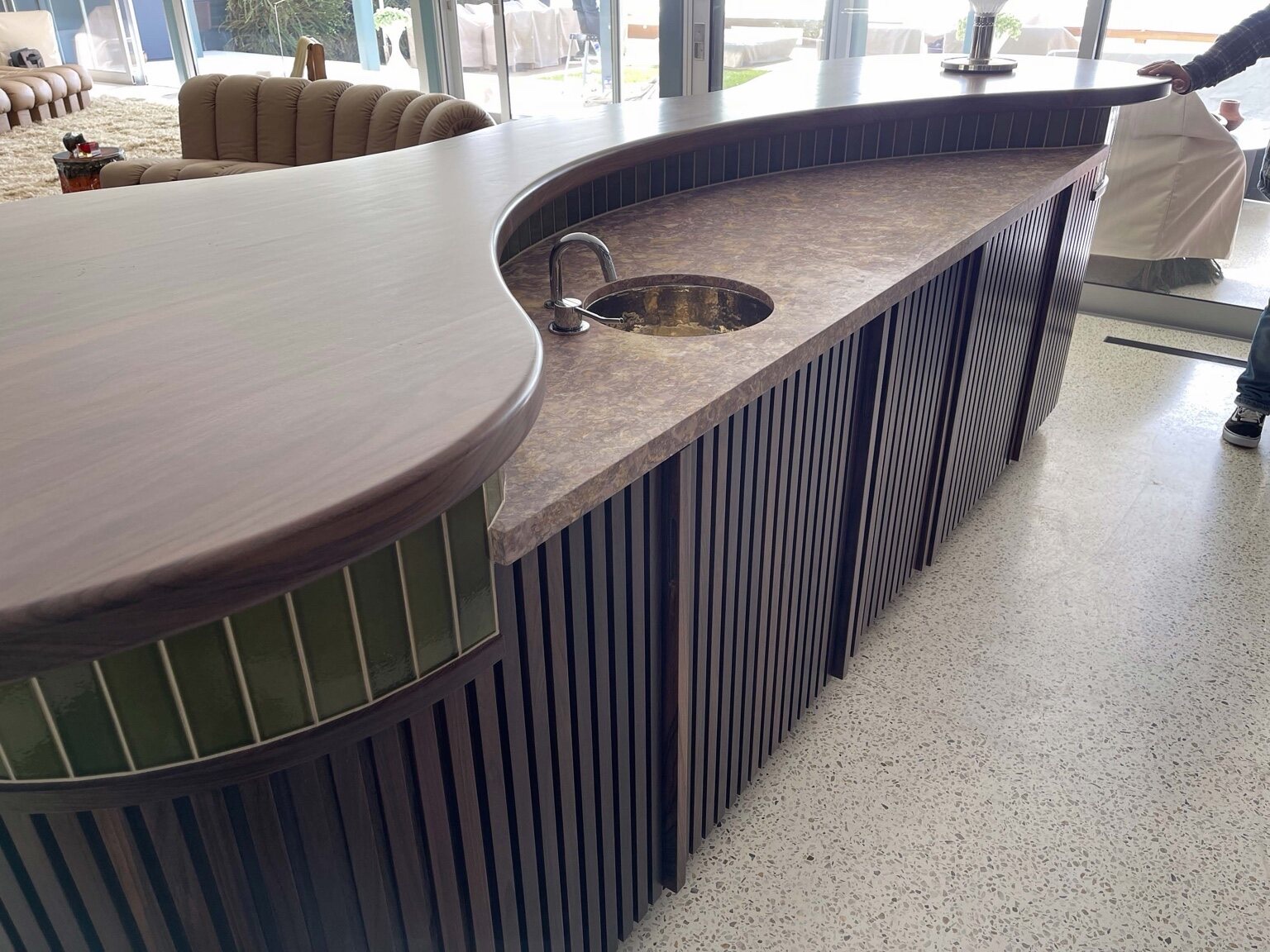 Kidney-shaped custom bar with a wood top and marbled stone inset, an undermount hammered-metal prep sink with a chrome gooseneck faucet, wood slat cladding, and a glossy green tile accent band