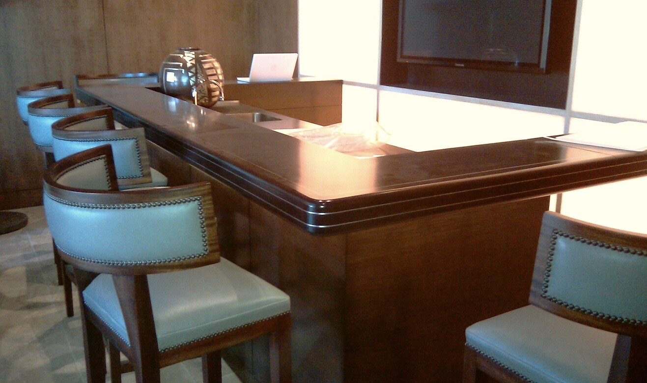 Traditional L-shaped custom wood bar with a molded ogee edge, matching bar-height stools upholstered in pale leather with nailhead trim, and backlit stone panels