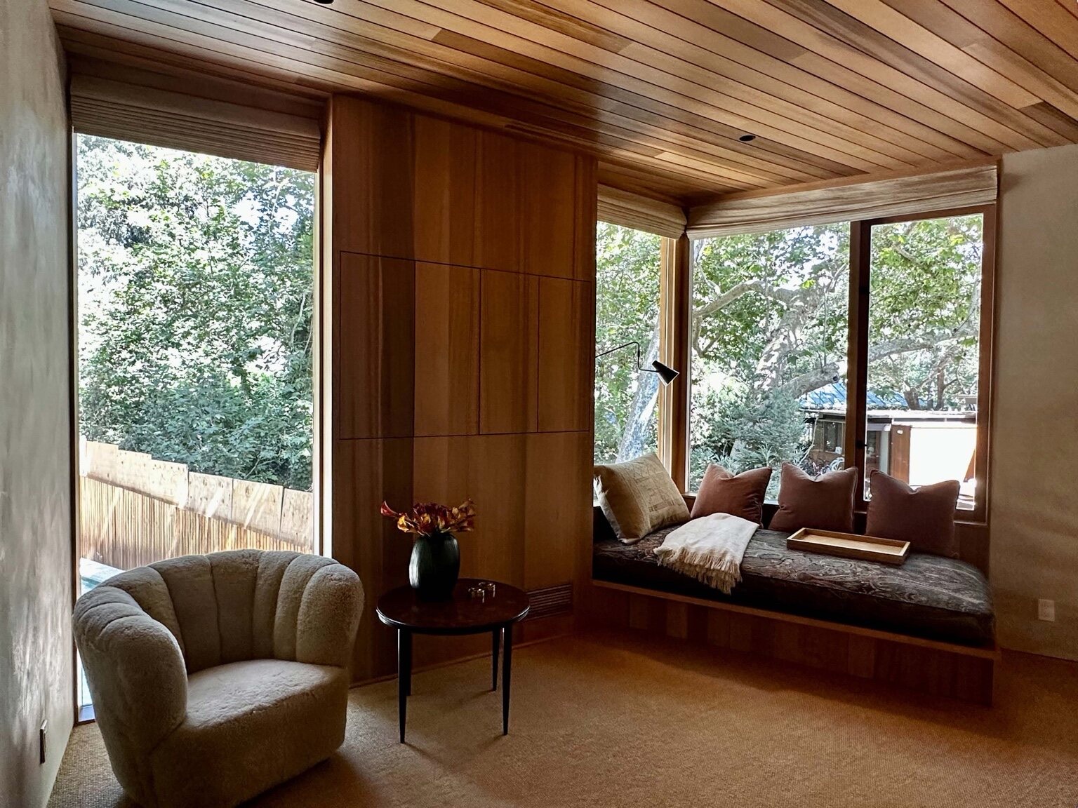 Warm wood-clad bedroom with a built-in window-seat daybed, wood-plank ceiling, and paneled wall overlooking trees
