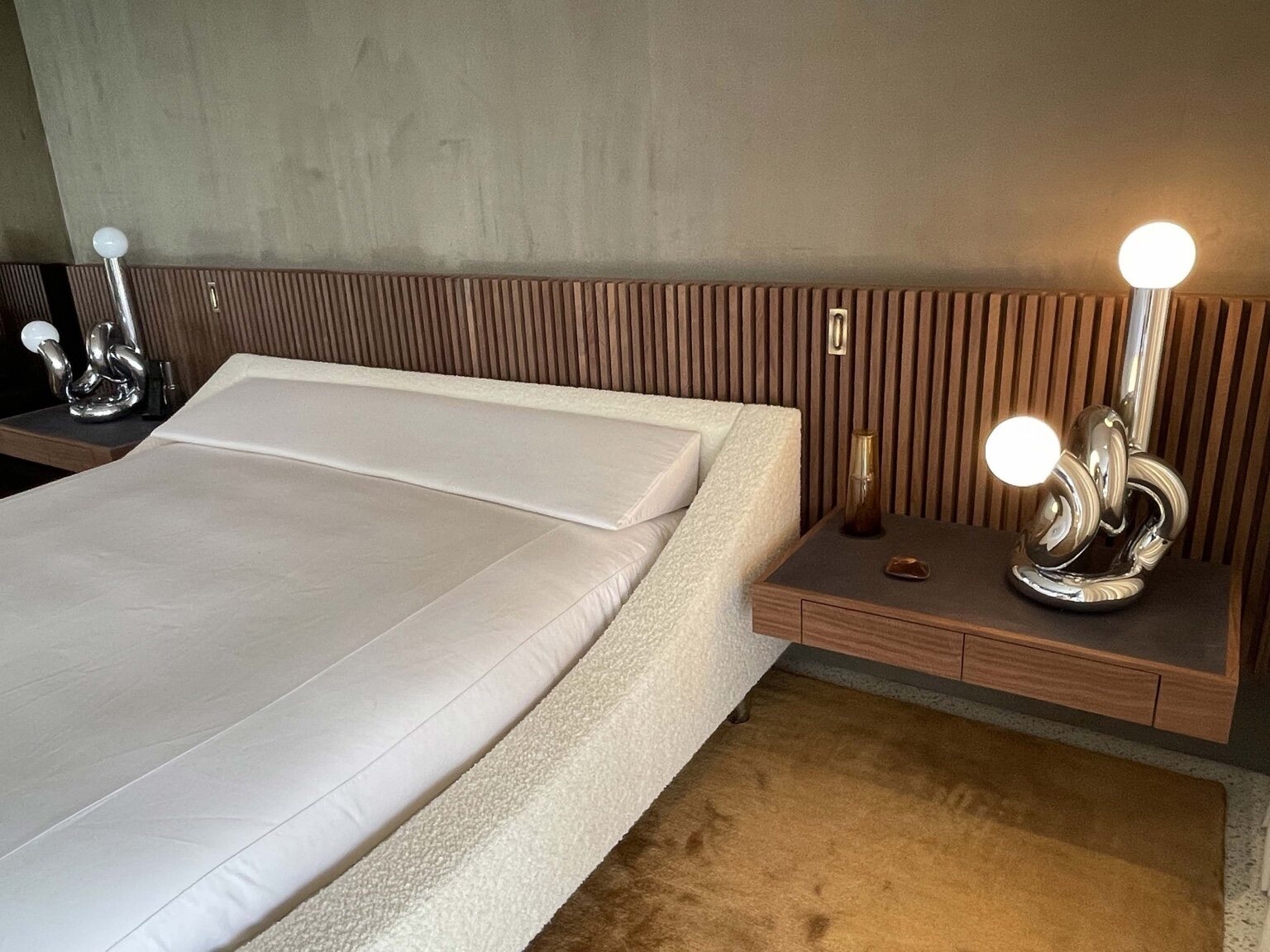 Modern bedroom with a low upholstered bouclé platform bed, a wood vertical-slat headboard wall with recessed pulls, and floating wood nightstands with chrome knot-form lamps
