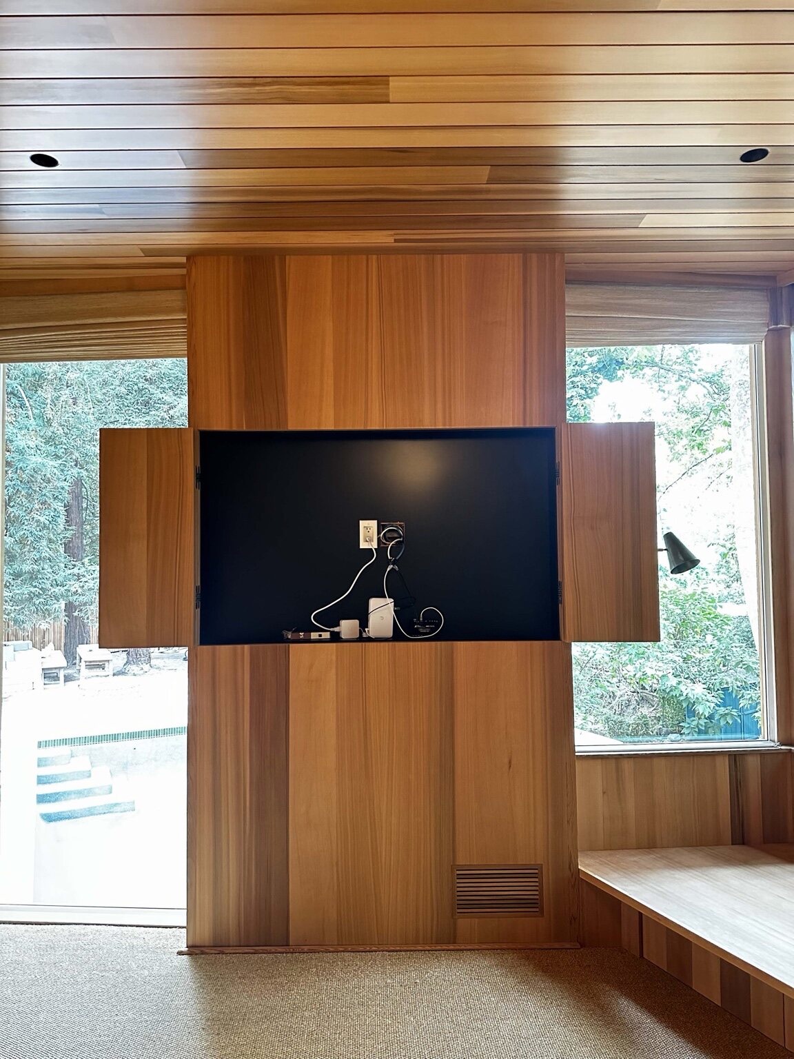 Wood-clad bedroom wall with a built-in media cabinet housing a TV in a recessed niche flanked by bi-fold wood doors, corner windows onto trees, and a built-in window bench