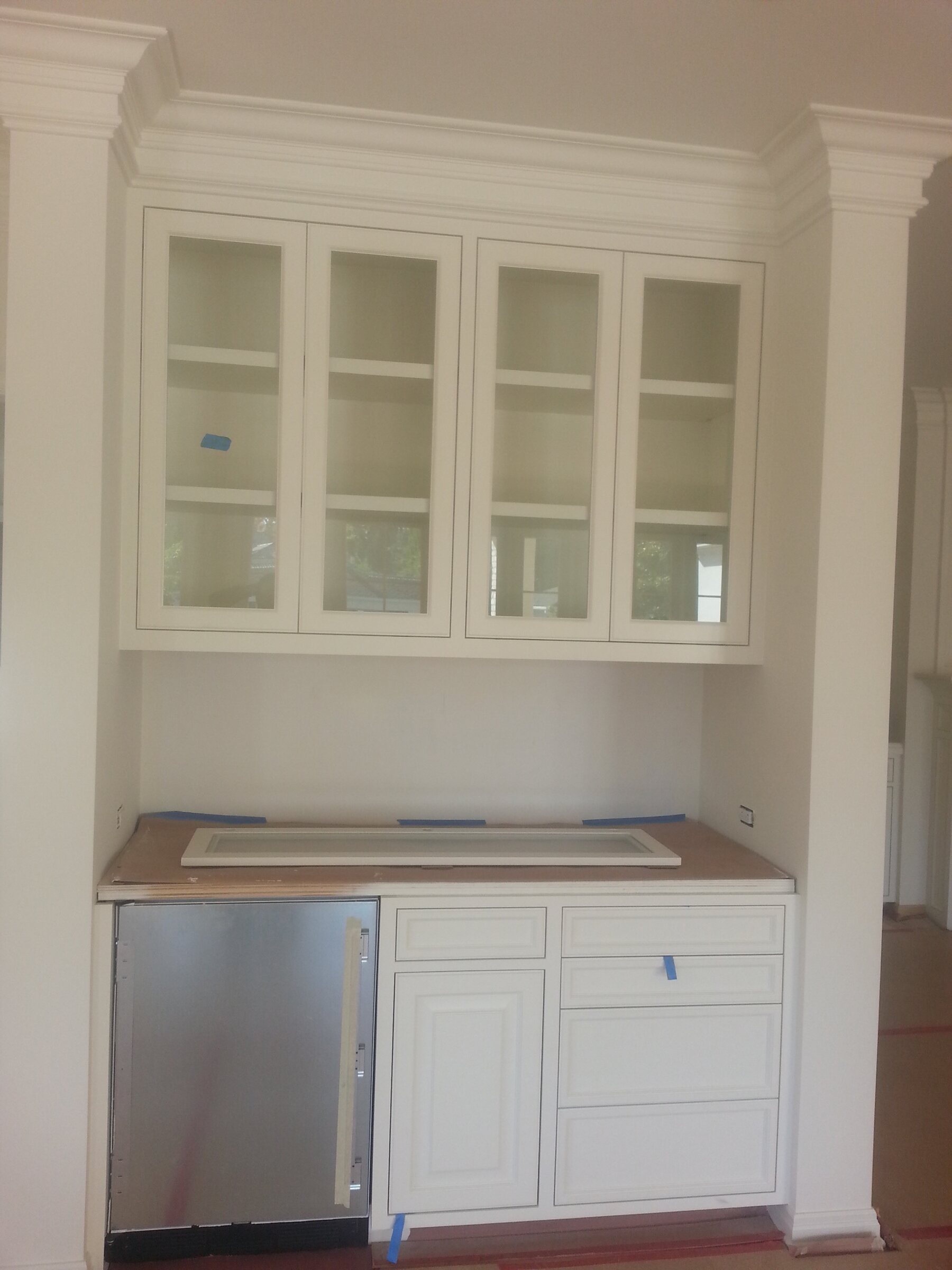 White painted butler's pantry beverage station with glass-front upper cabinets, an under-counter stainless refrigerator, and raised-panel base cabinets in progress