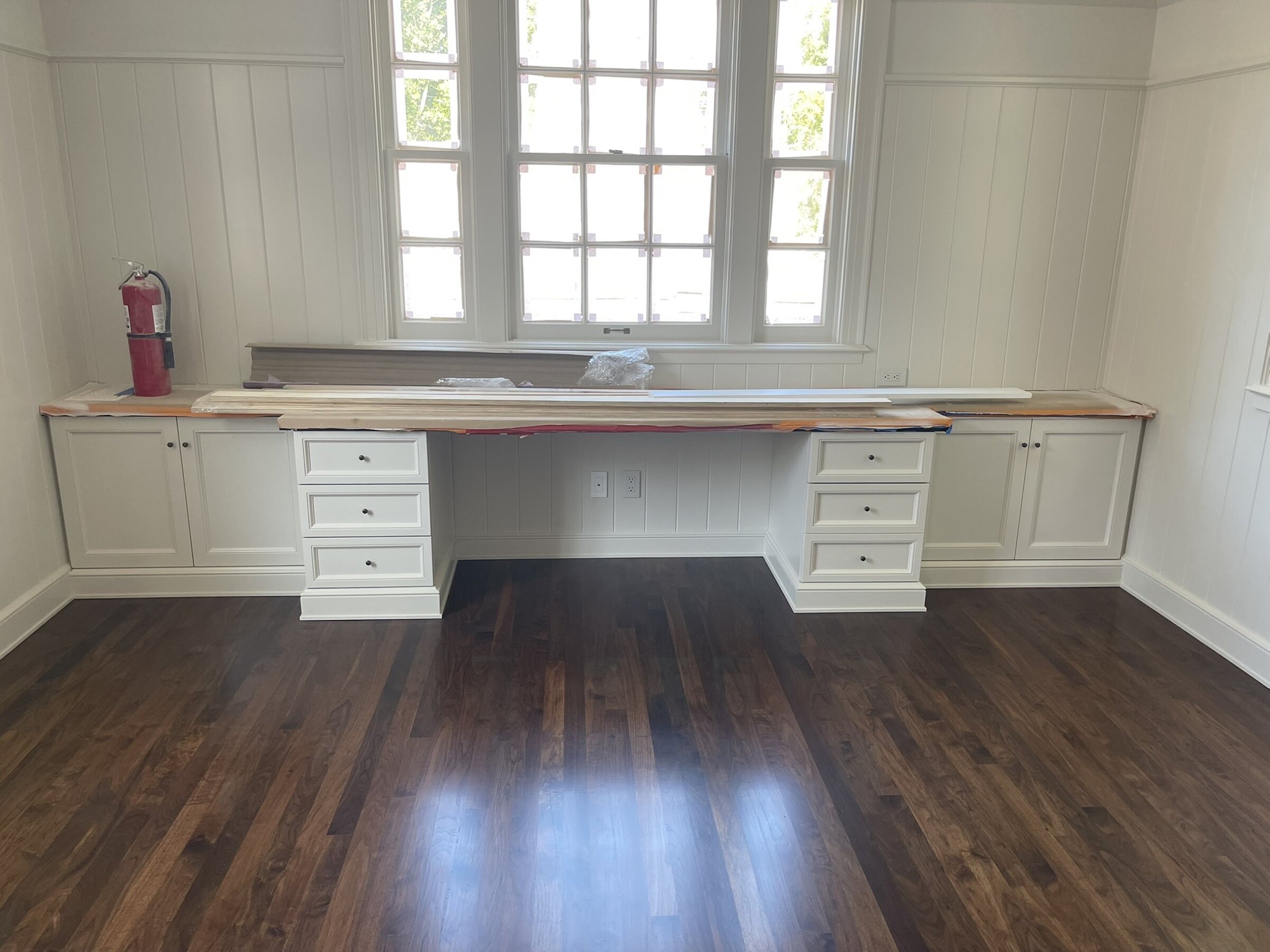 White painted built-in home office with a long desk between two three-drawer pedestals, flanking base and wall cabinets, board-and-batten paneling, and dark wood floors
