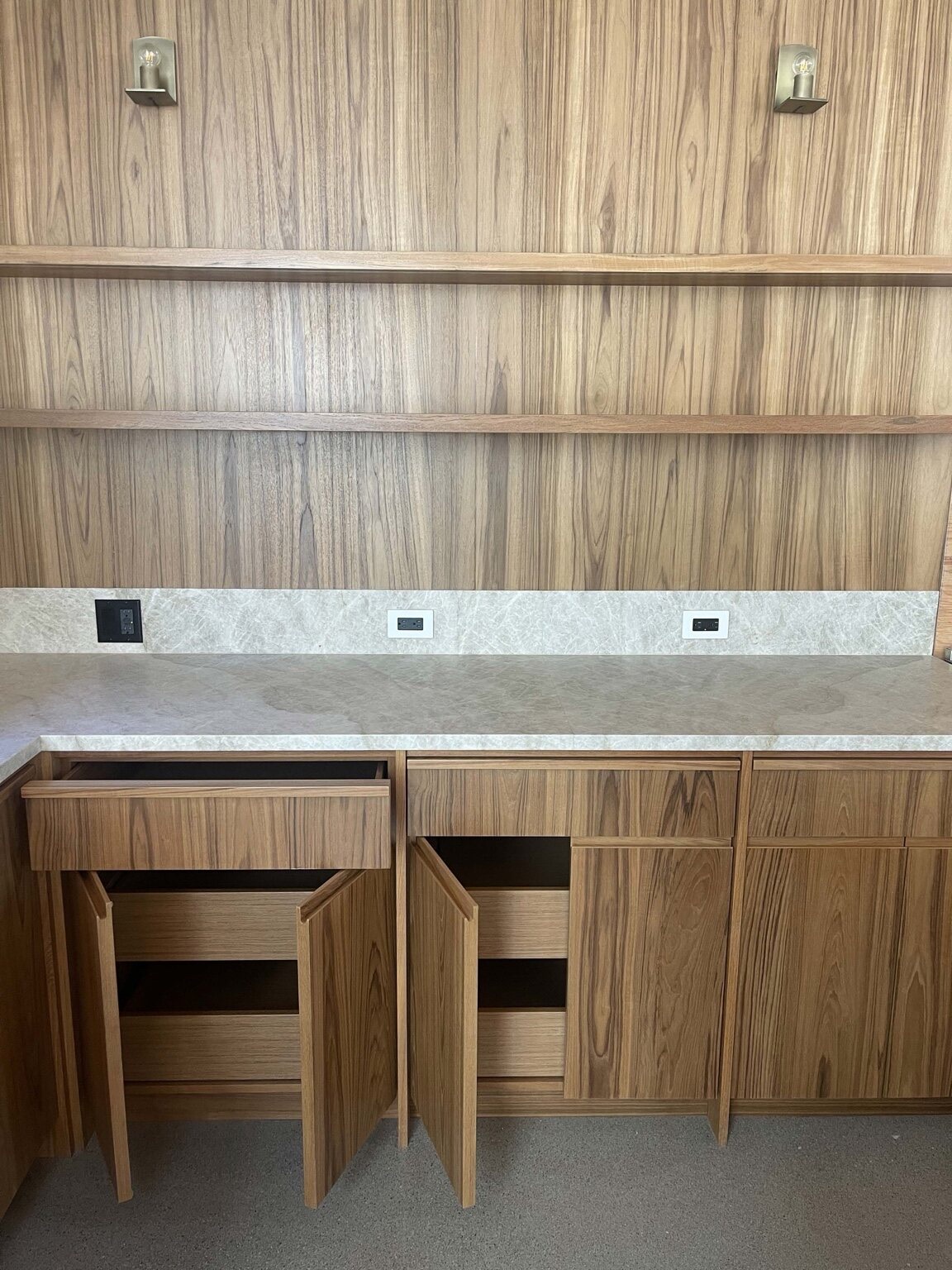Modern vertical-grain wood cabinetry with a stone counter and backsplash, floating shelves, and wall sconces, with a drawer and doors open