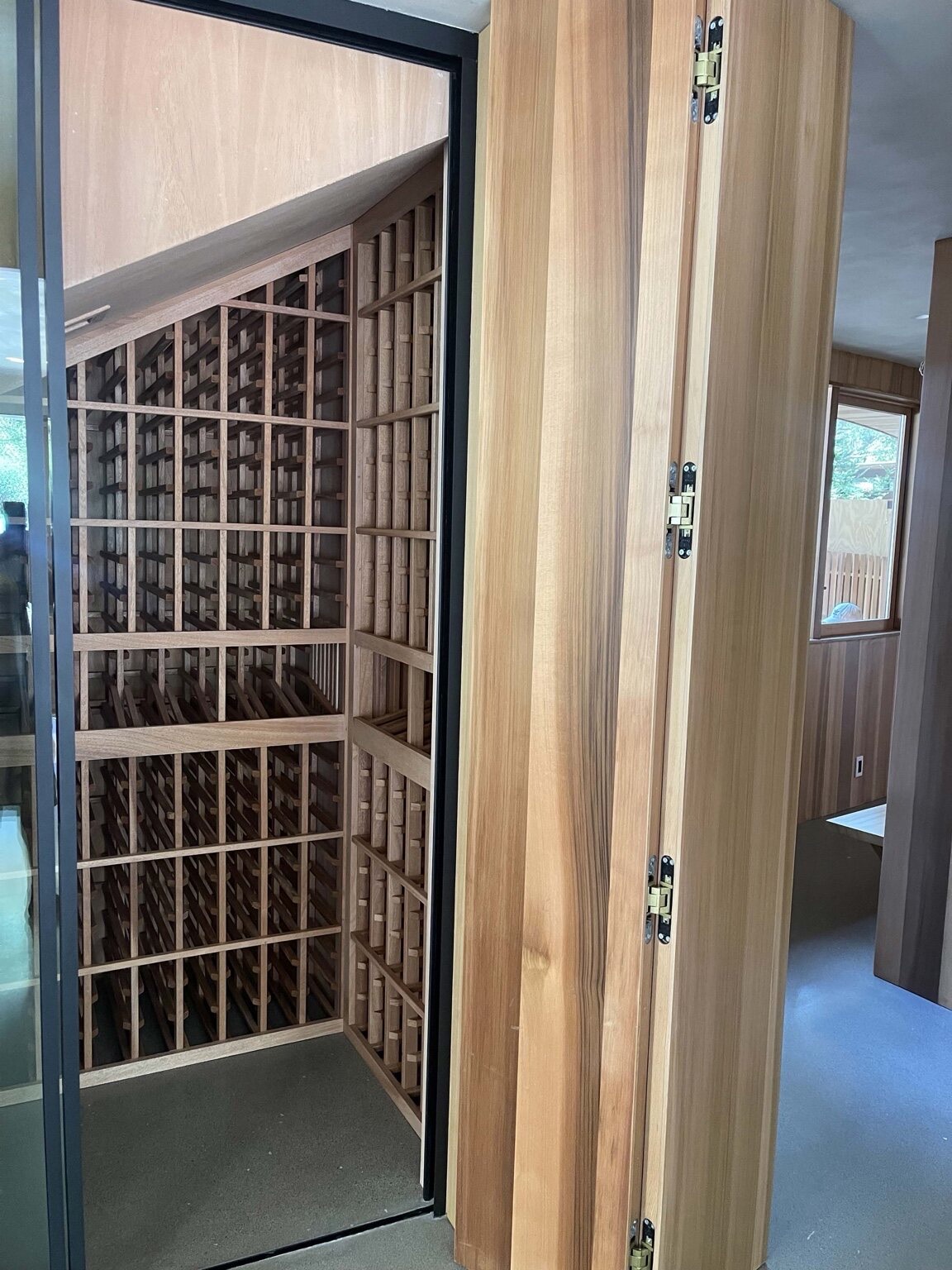 Custom wood wine cellar with diamond-bin and column bottle racking behind a glass door, beside a tall wood pivot door with concealed hinges