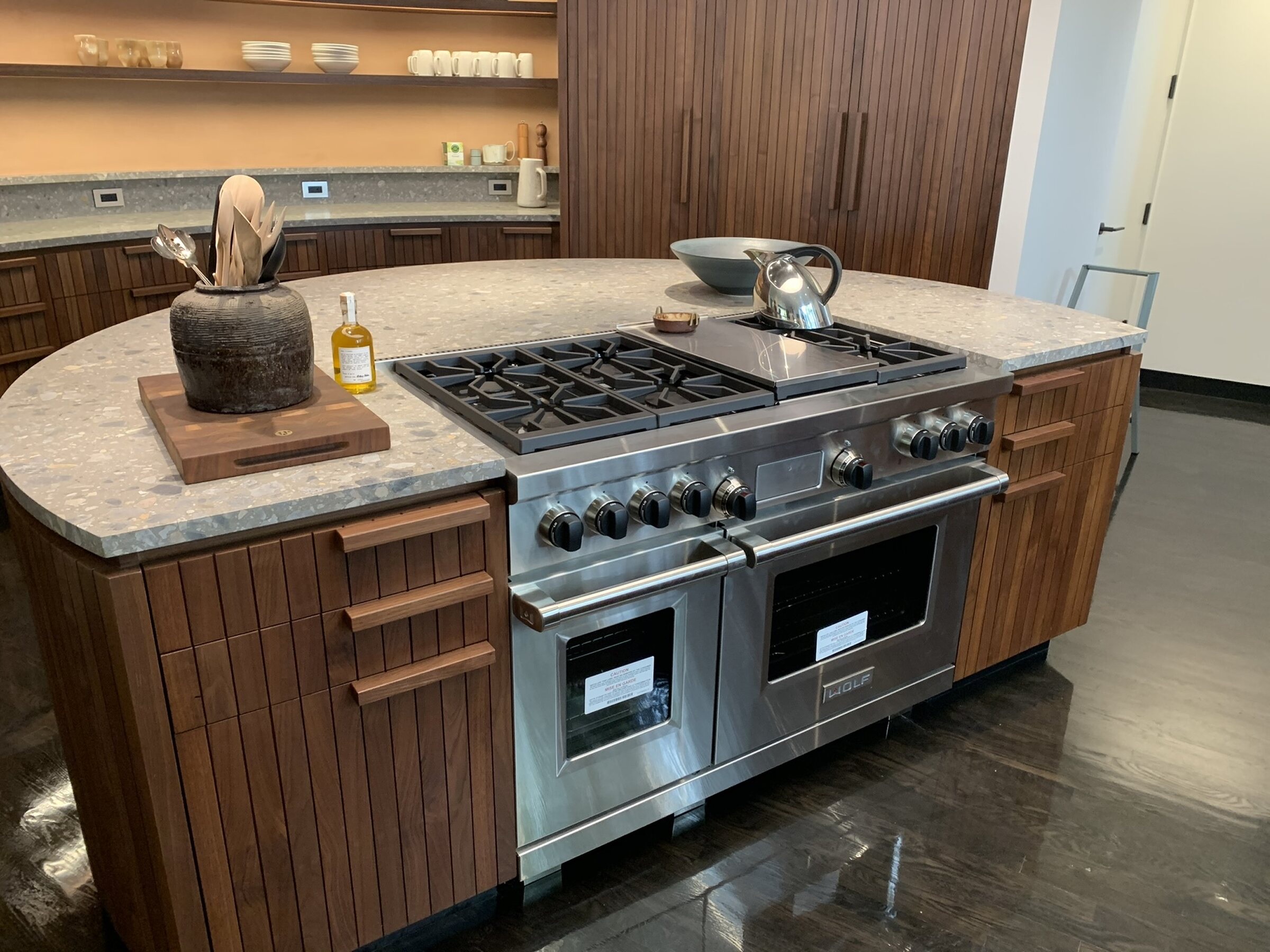 Curved stone island housing a large stainless range, flanked by slatted wood drawer banks with lit open shelving behind