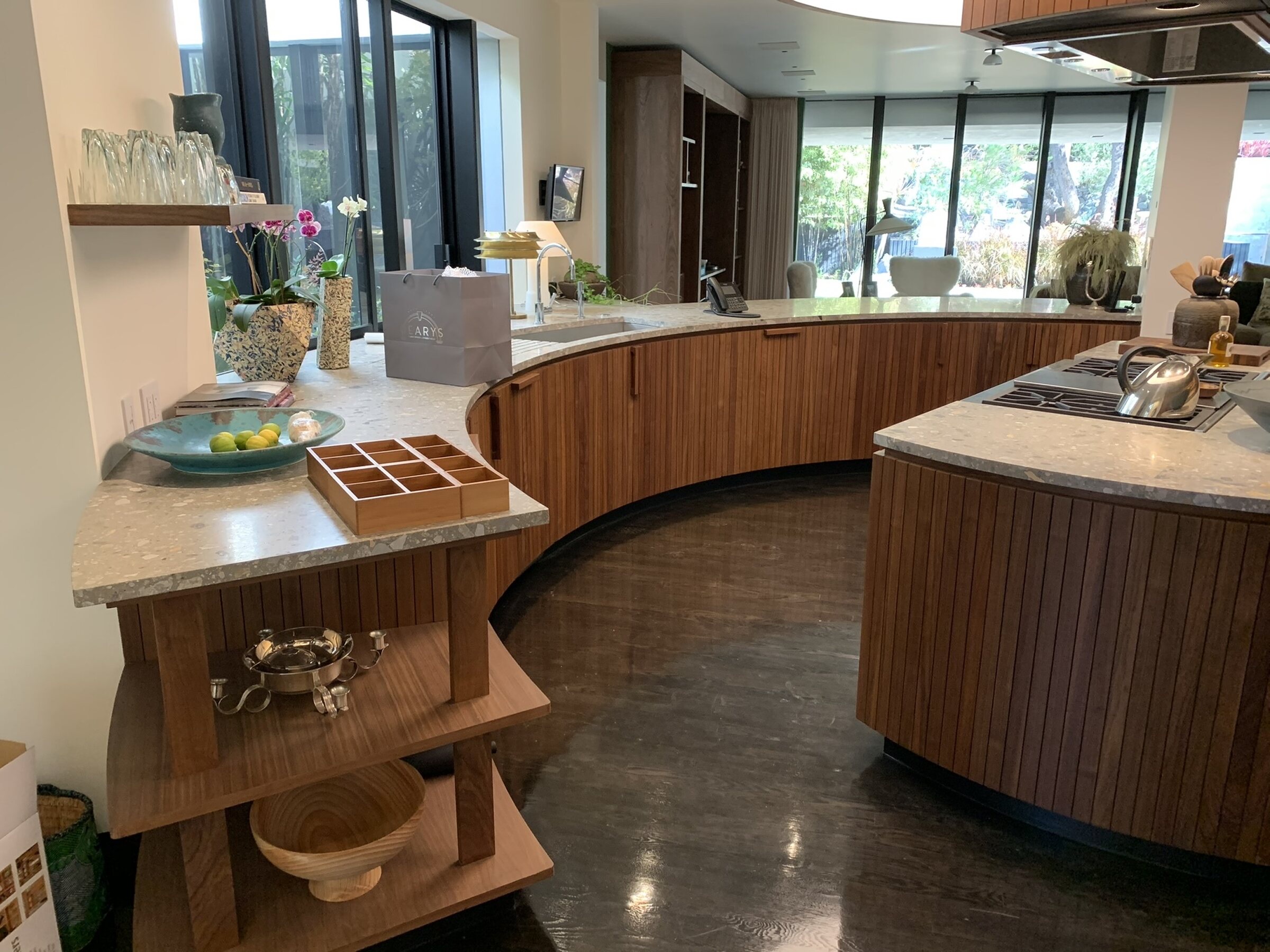 Wide view of a sweeping curved run of slatted wood cabinetry with stone countertops, a curved island with a bar stool, and dark polished floors