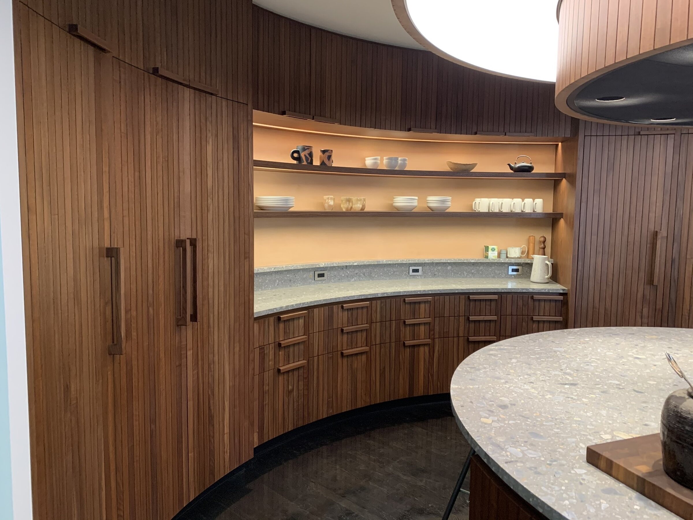 Curved vertical-slat wood cabinetry wrapping a wall with backlit open shelving of dishware, a peach accent wall, and stone counters