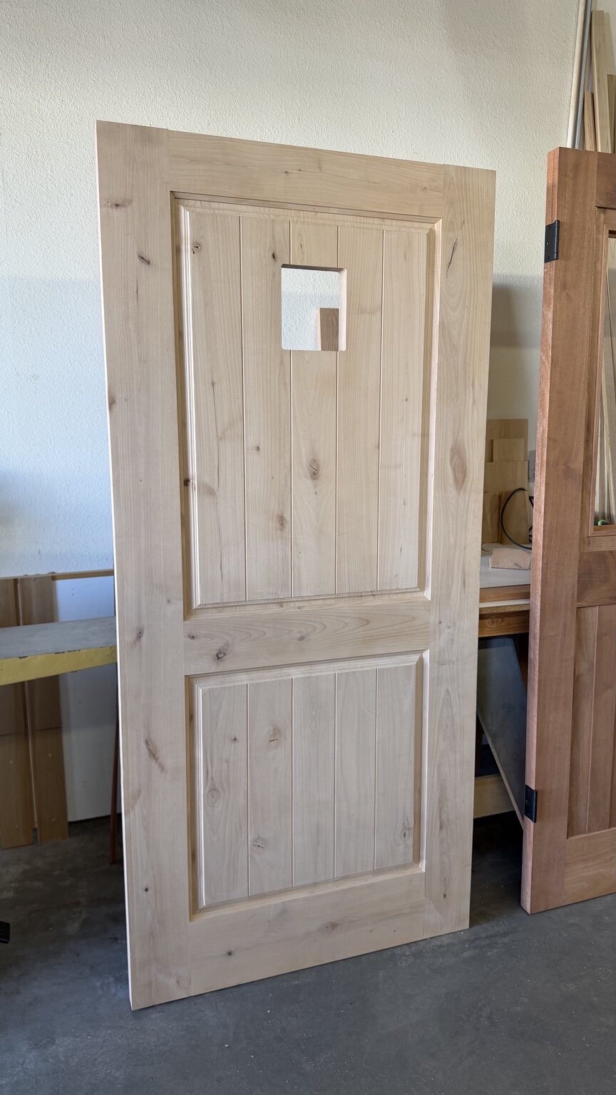 Rustic custom wood two-panel door with V-groove plank panels and a cut-out for a pet or speakeasy opening, unfinished in the shop
