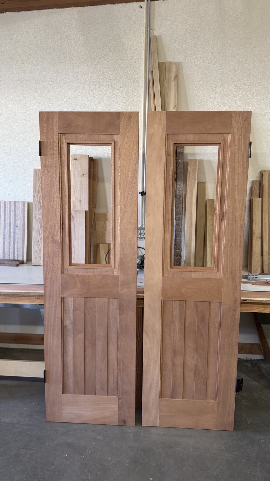 Pair of custom wood doors with upper glass lights and lower V-groove plank panels, standing unfinished in the workshop
