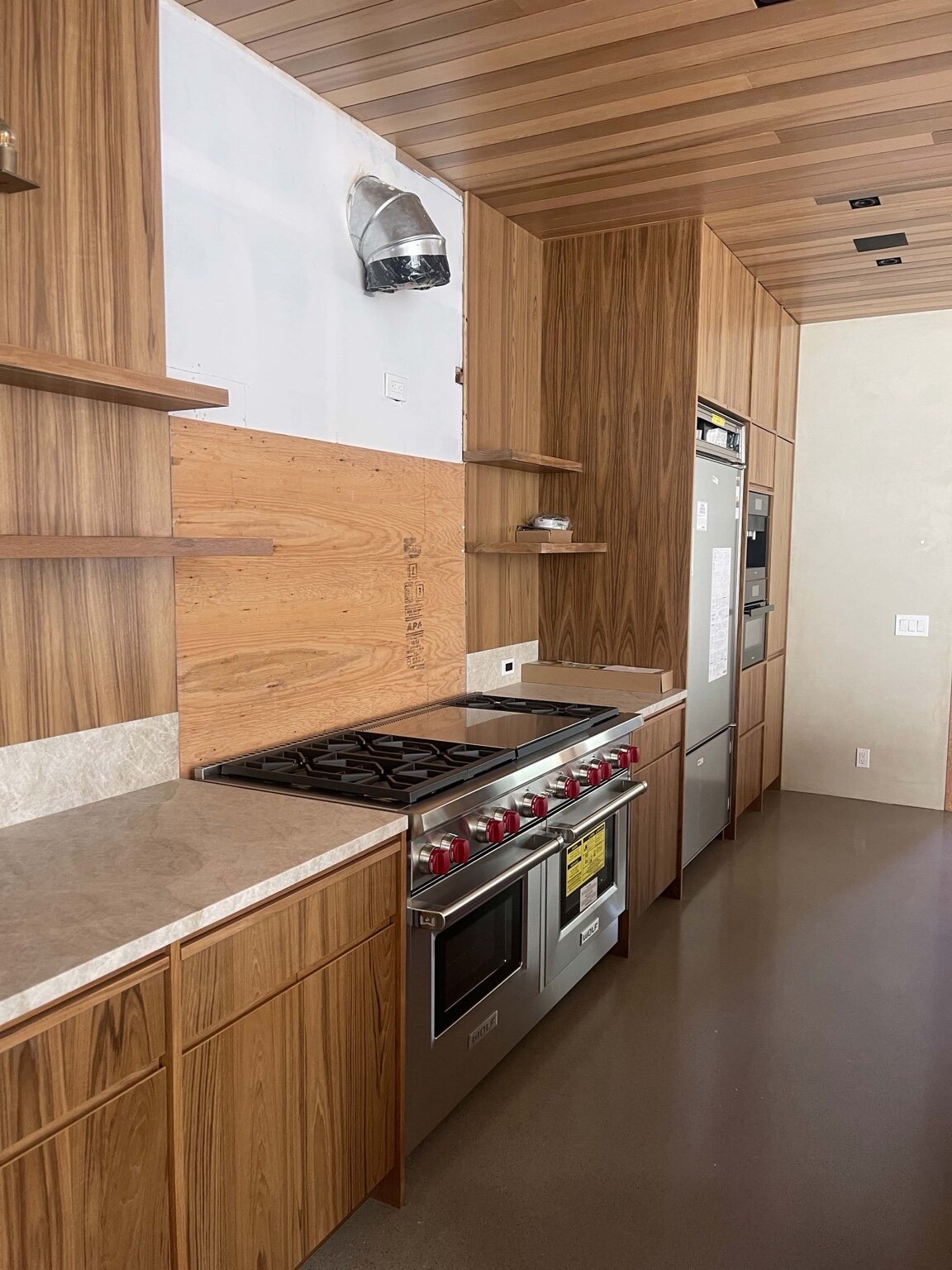 Galley kitchen during installation with a stainless professional range, wood-slat ceiling, floating shelves, and an integrated tall refrigerator