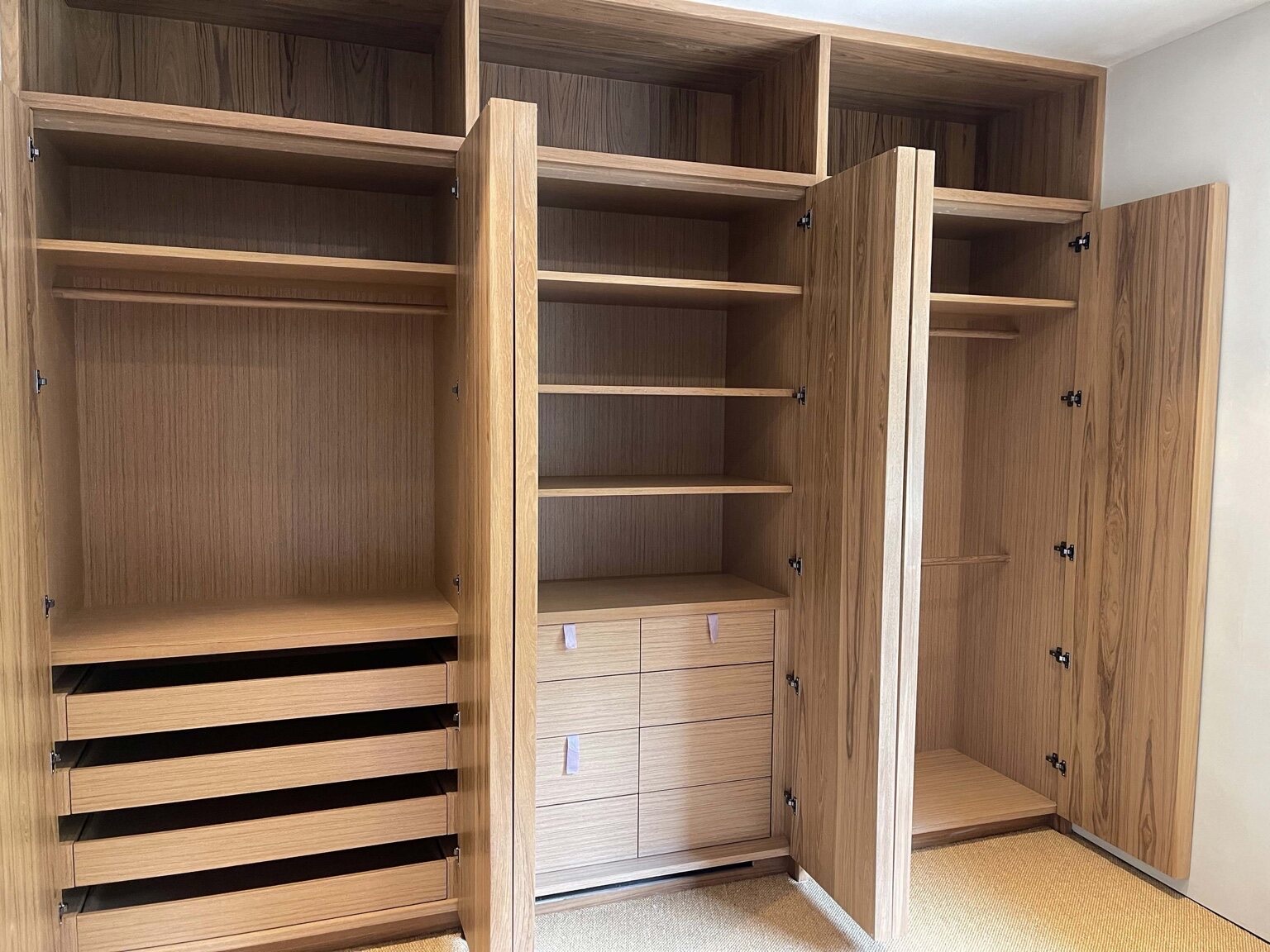 Interior of a custom wood fitted wardrobe with bi-fold doors open, revealing adjustable shelves, hanging rods, and pull-out drawers