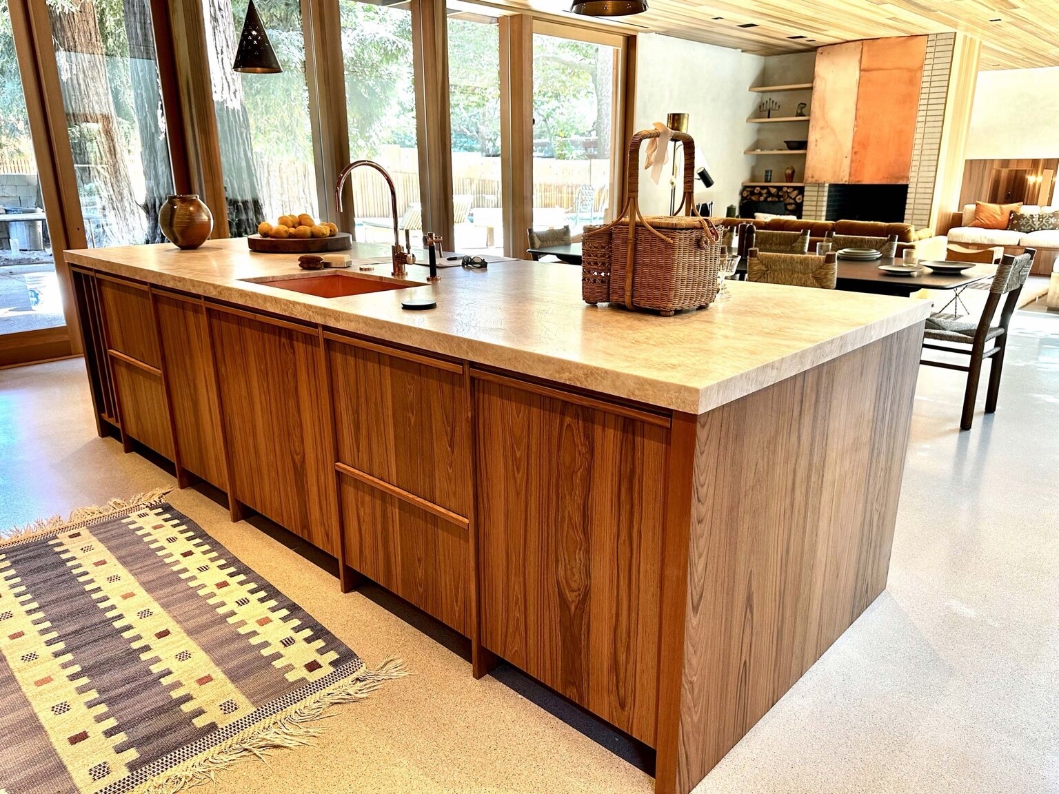 Long custom wood kitchen island with a waterfall stone countertop and integrated sink, set in an open-plan great room with floor-to-ceiling glass