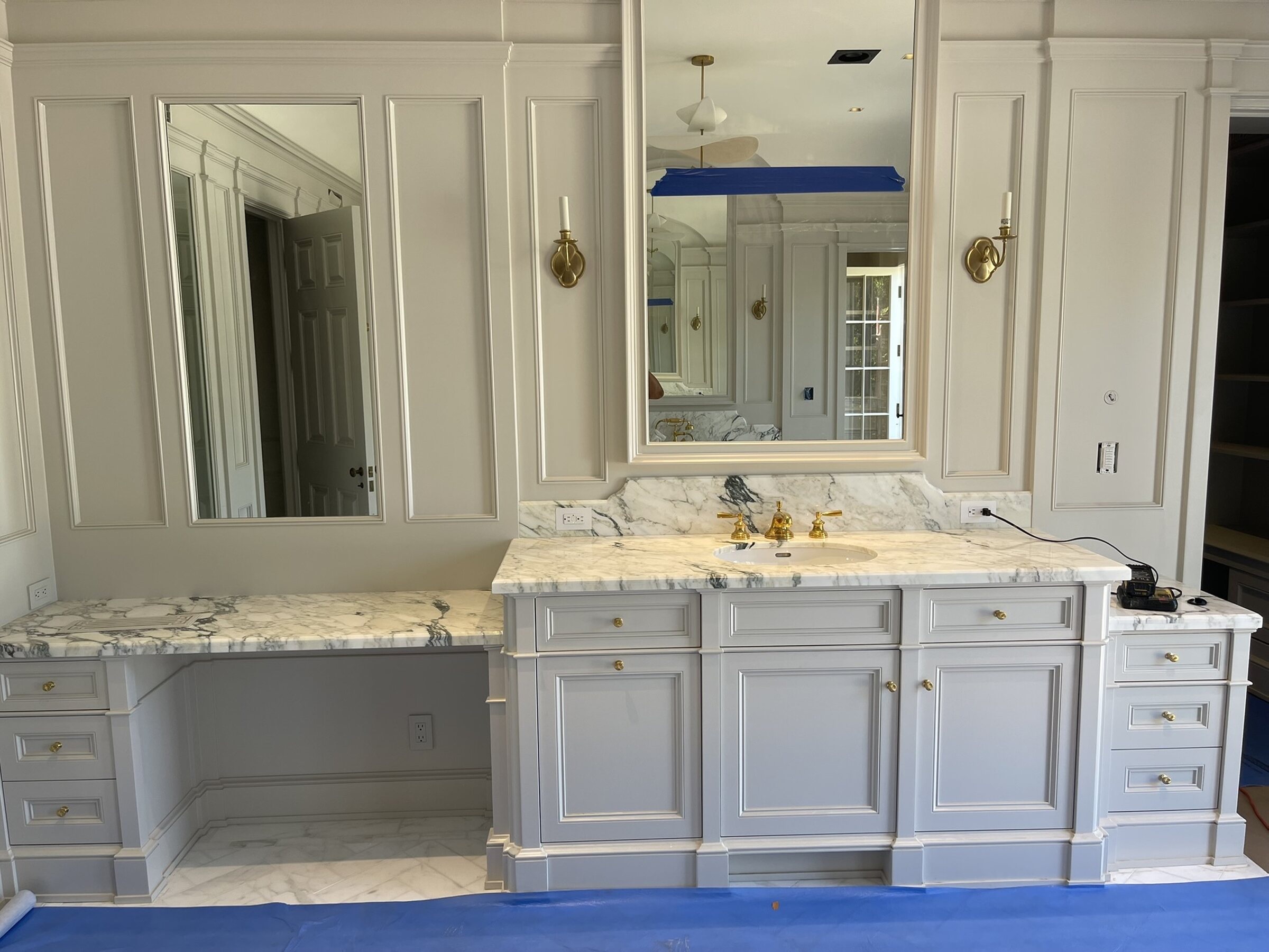 Large painted master bathroom vanity with marble counters, gold fixtures, framed mirrors, and detailed paneled millwork