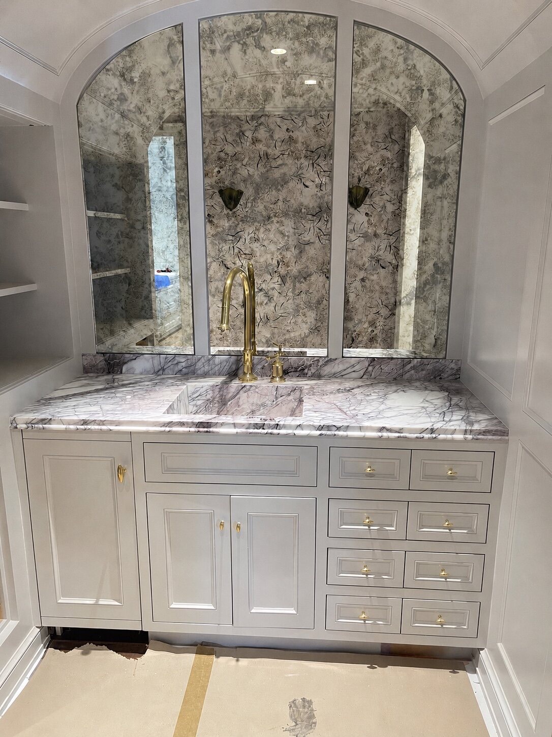 Light grey painted inset vanity with arched antiqued mirror panels, veined lilac-white marble counter, undermount sink, and brass gooseneck faucet with teardrop knobs