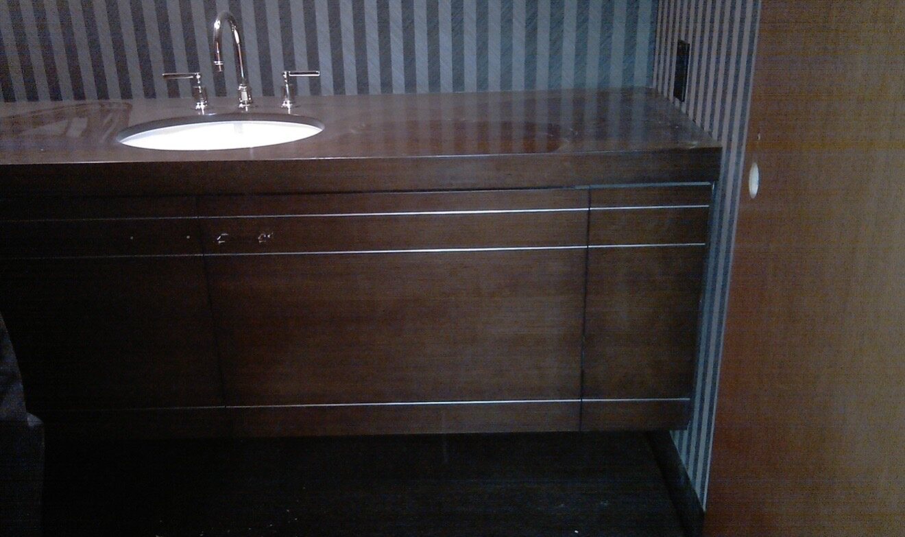 Dark custom-stained vanity with fine reveal grooves, a polished dark stone top, undermount oval sink, and chrome gooseneck widespread faucet against striped wallpaper