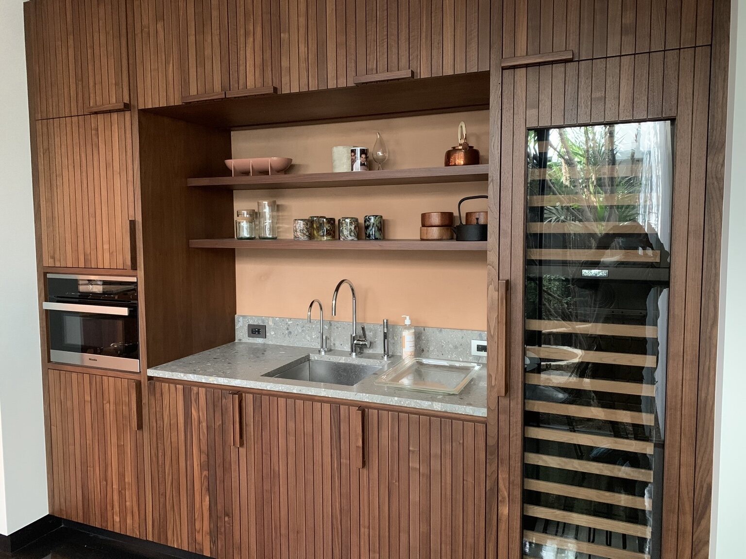 Wood slat kitchenette with undermount sink, stone counter, floating shelves, a built-in oven, and a glass-front wine refrigerator
