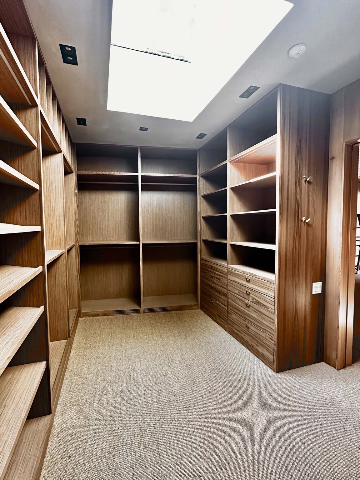 Finished custom wood walk-in closet with floor-to-ceiling open shelving, hanging sections, a bank of drawers, and a skylight above beige carpet